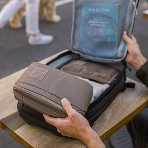 Person packing a CIVIC Access Pouch 2L in Fossil Brown - travel organizer into an open suitcase on a wooden table.