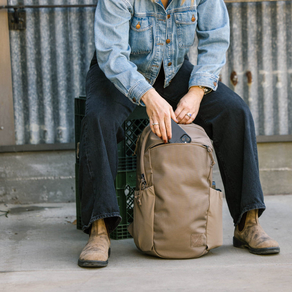 Person sitting on a crate with a CIVIC Half Zip 22L in Fossil Brown backpack, wearing a denim jacket and dark pants. Person sitting on a crate with a CIVIC Half Zip 22L in Fossil Brown backpack, wearing a denim jacket and dark pants. [13003d-FBN