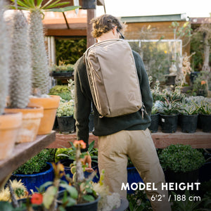 Person with a CIVIC Panel Loader 24L in Fossil Brown backpack standing in a garden center surrounded by plants.