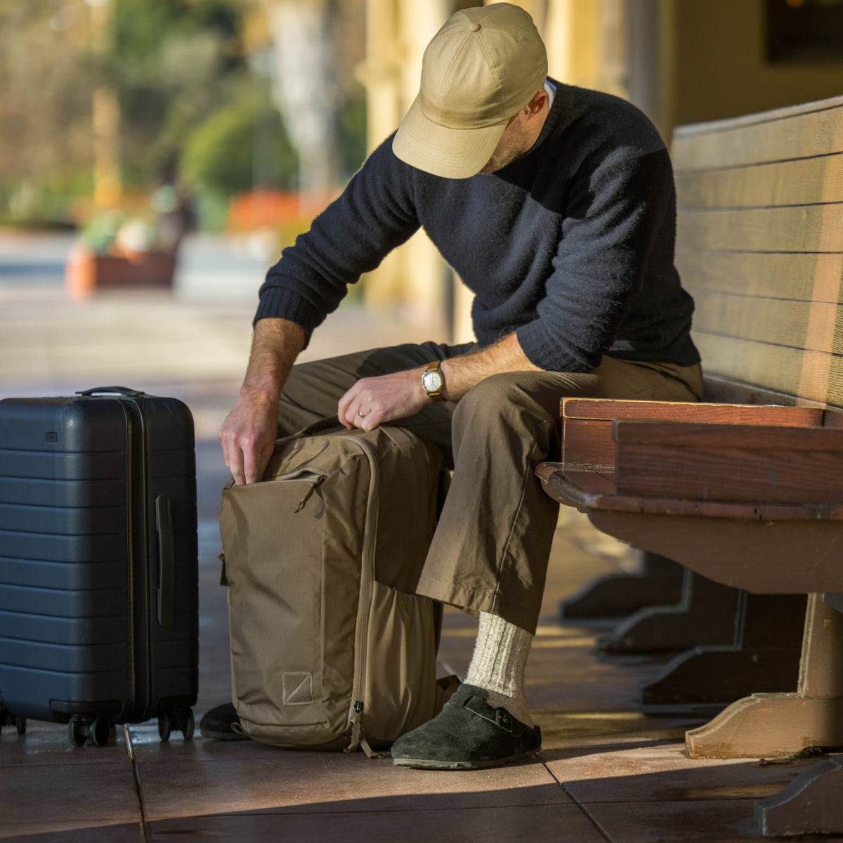 Man sitting on a bench with a CIVIC Travel Bag 26L in Fossil Brown backpack and suitcase accessing front dump quick access pocket. Man sitting on a bench with a CIVIC Travel Bag 26L in Fossil Brown backpack and suitcase accessing front dump quick access pocket. [13012c-FBN