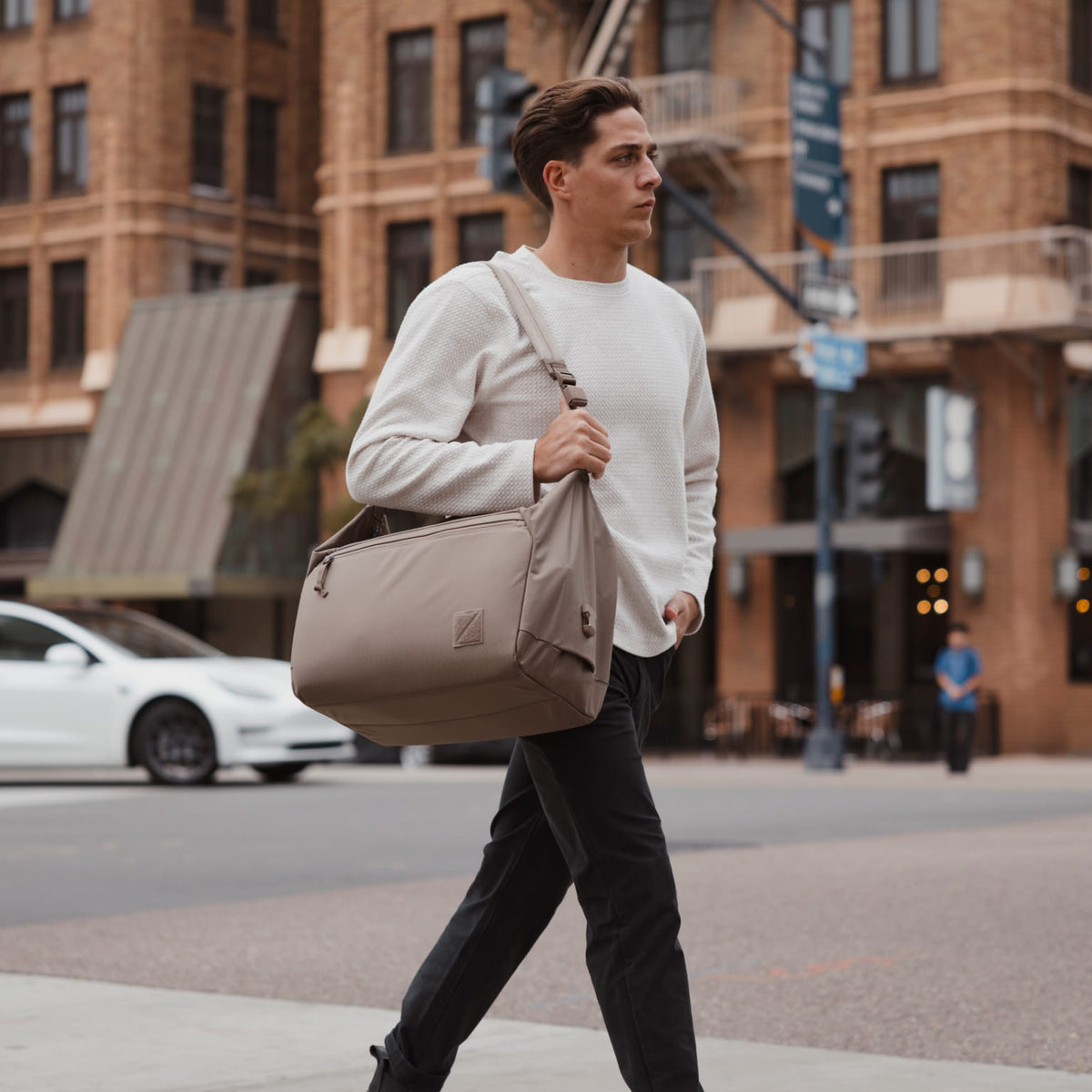 A man in a white sweater and black pants walks across a city street while carrying a large taupe-beige duffel bag over his shoulder. A man in a white sweater and black pants walks across a city street while carrying a large taupe-beige duffel bag over his shoulder. [13023d-FBN