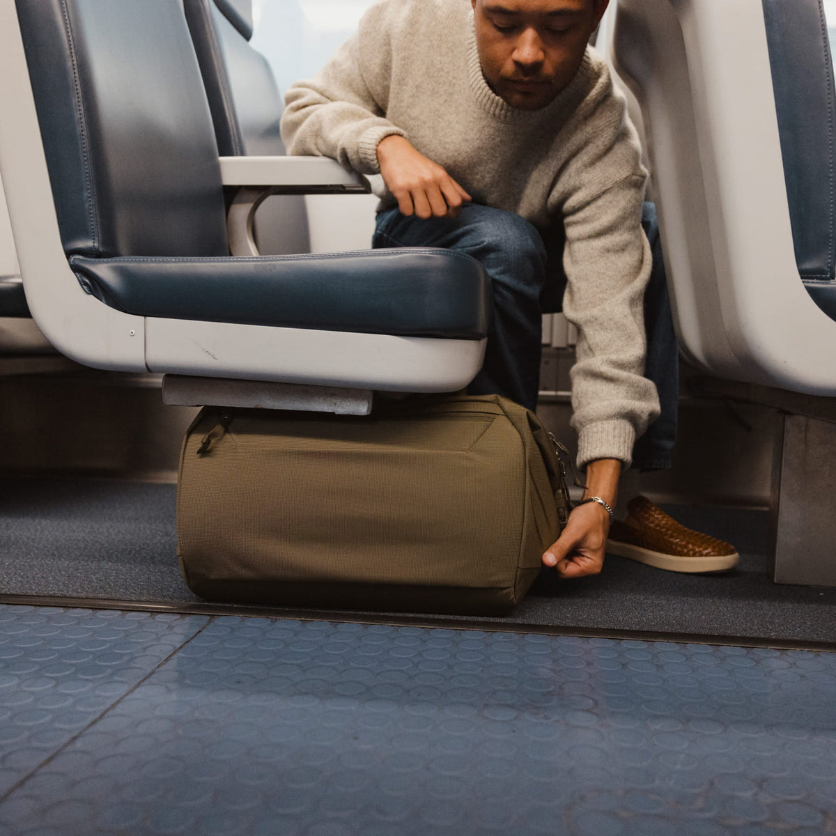 Man adjusting a Transit Duffel 25L in OD Green under an airplane seat Man adjusting a Transit Duffel 25L in OD Green under an airplane seat [13023d-ODG