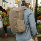 Person wearing a CIVIC Half Zip 22L in Fossil Brown backpack in an outdoor market with people and plants. Person wearing a CIVIC Half Zip 22L in Fossil Brown backpack in an outdoor market with people and plants. [13003d-FBN