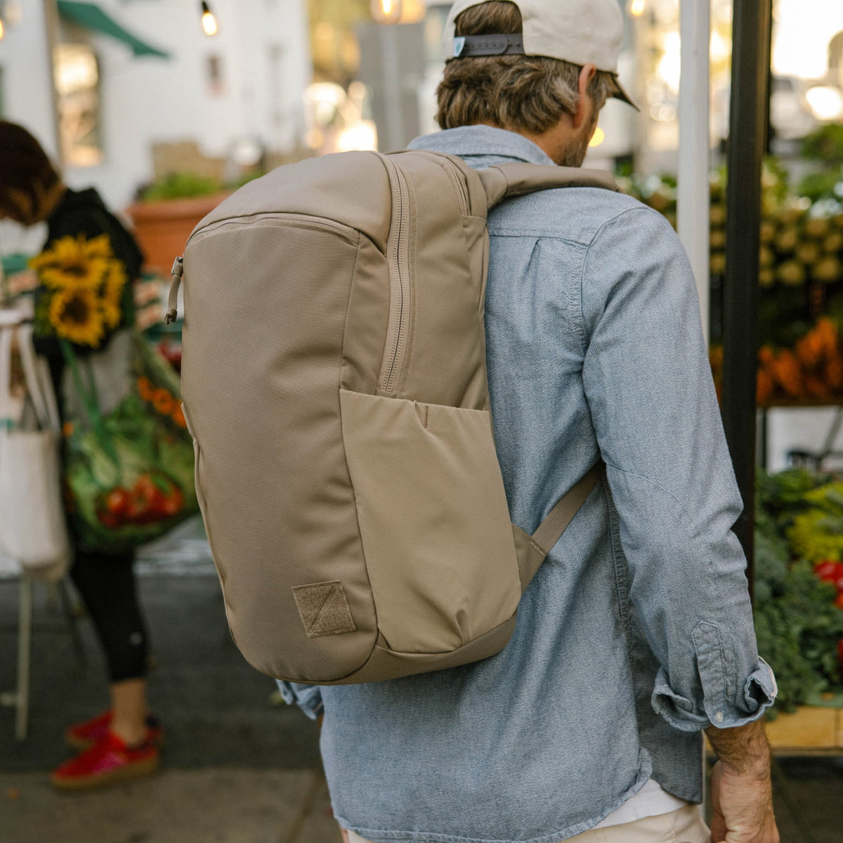 Person wearing a CIVIC Half Zip 22L in Fossil Brown backpack in an outdoor market with people and plants. [13003d-FBN]
