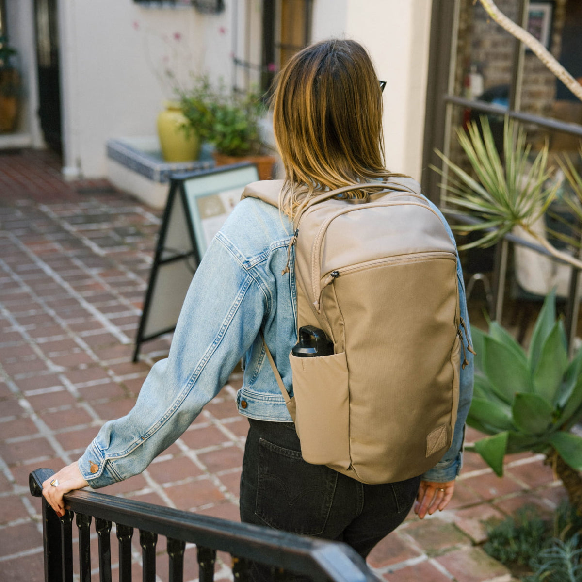 Person wearing a CIVIC Half Zip 22L in Fossil Brown backpack in an outdoor setting with plants and a building in the background [13003d-FBN]