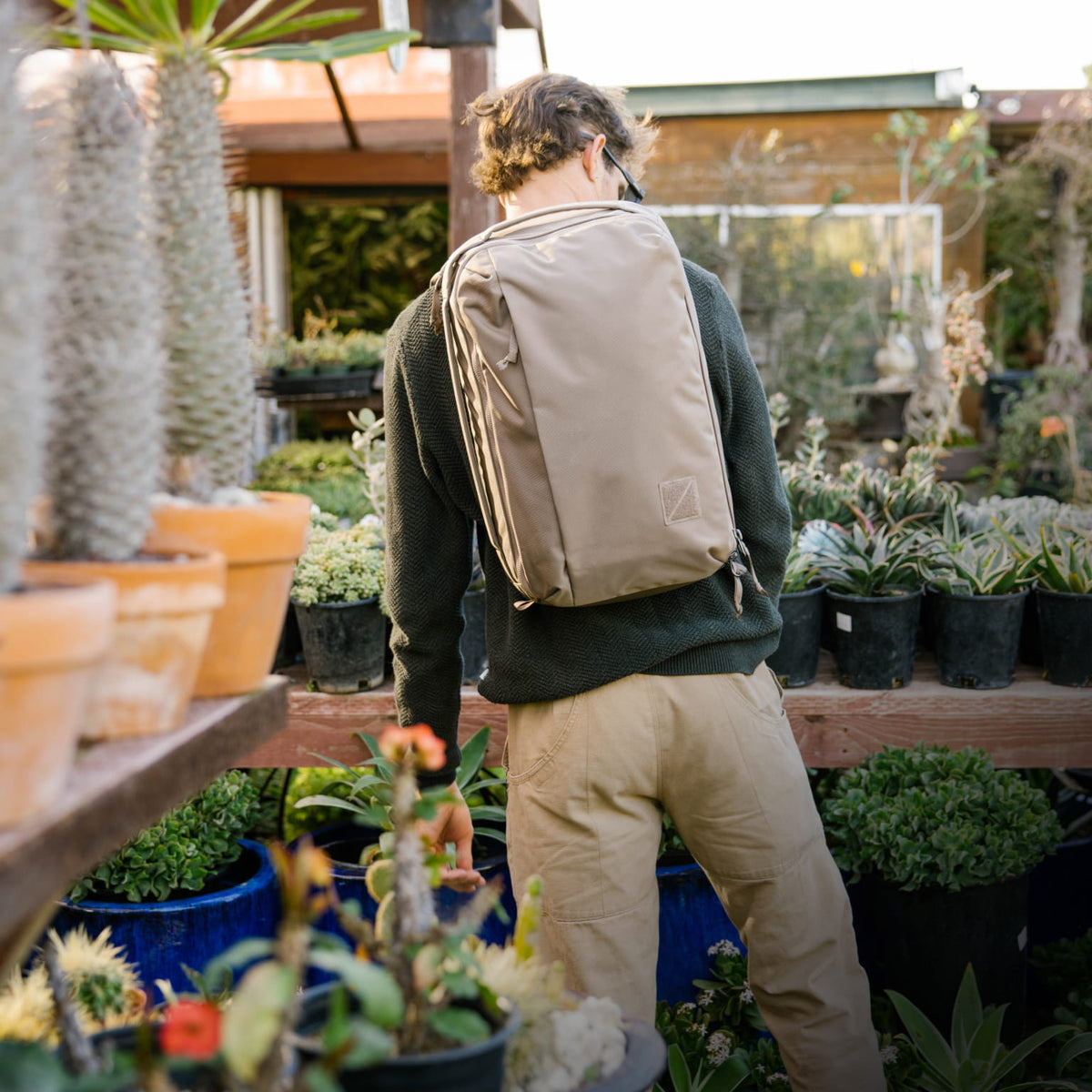Person with a CIVIC Panel Loader 24L in Fossil Brown  backpack standing in a garden center surrounded by plants. [13001c-FBN]