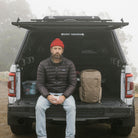 Man sitting in the back of a vehicle with a backpack, wearing a red beanie and dark jacket. CIVIC Travel Bag 20L in Fossil Brown. Man sitting in the back of a vehicle with a backpack, wearing a red beanie and dark jacket. CIVIC Travel Bag 20L in Fossil Brown. [13019c-FBN