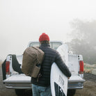 Person with a CIVIC Travel Bag 20L in Fossil Brown and surfboard standing next to a white truck in a foggy landscape. Person with a CIVIC Travel Bag 20L in Fossil Brown and surfboard standing next to a white truck in a foggy landscape. [13019c-FBN