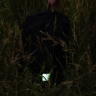 A black backpack lying in tall grass, with a hand holding the top handle and a small glowing patch attached near the bottom front.